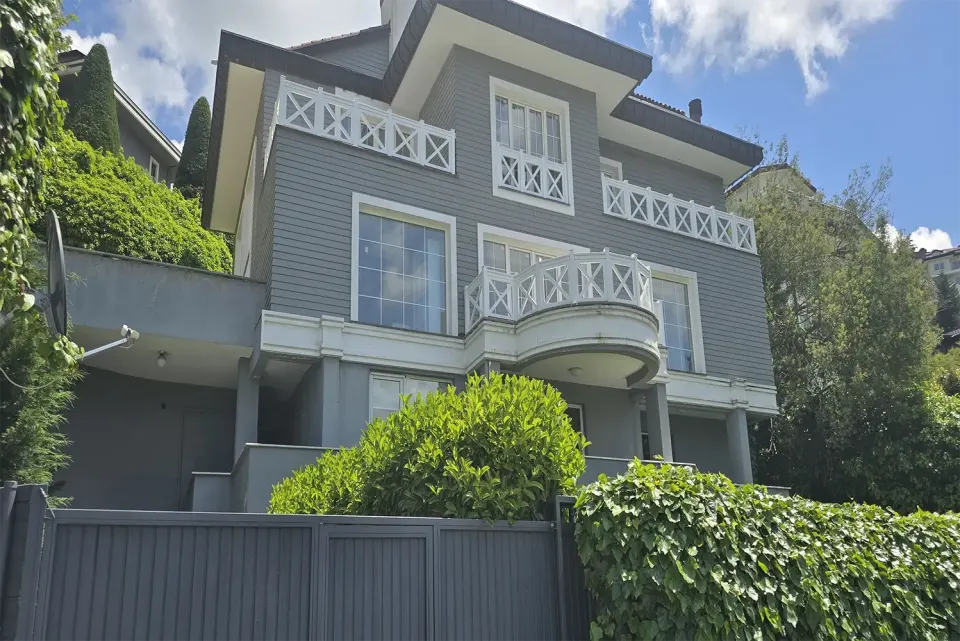 Yarım daire balkonlu gri villa cephesi ve önündeki sarmaşıklı bahçe duvarı