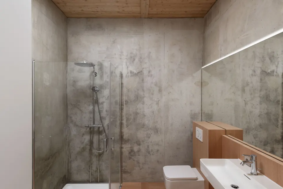 Ahşap villa banyosunda brüt beton duvarlar, cam duşakabin ve ahşap kaplı lavabo dolabı